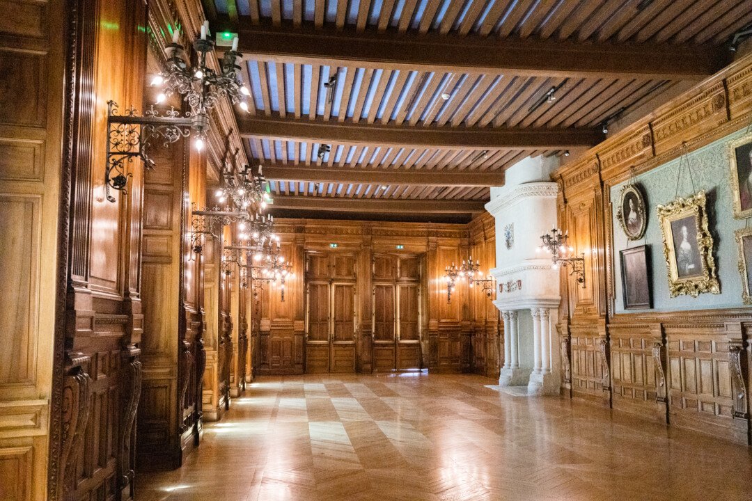 Salle du château de Grignan. Les murs sont couverts de bois et papier peint, avec quelques peintures accrochée. Des chandeliers en fer forgé sont également fixés à ces derniers à intervalles réguliers. On aperçoit dans le fond une massive cheminée en pierre claire.