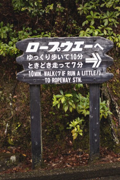 Un panneau de bois indique le chemin vers la station de téléphérique. Le texte (en japonais et anglais) dit : "10 minutes de marche jusqu'à la station de téléphérique (7 en courant un peu)"