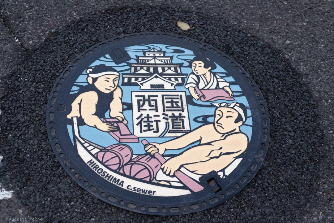 Plaque d'égout à Hiroshima présentant une illustration du château et divers personnages travaillant dans l'artisanat