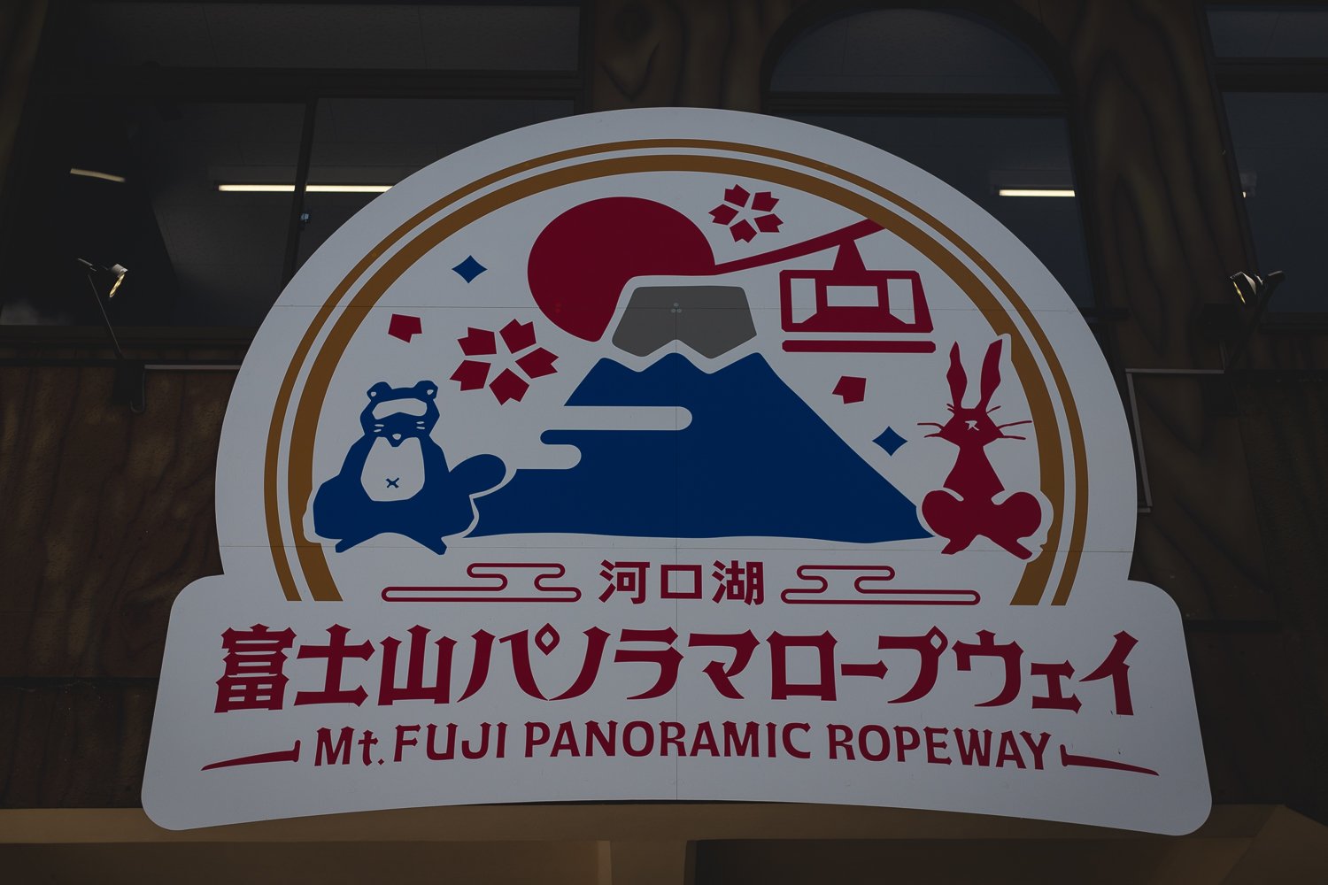 Signe à l'entrée du téléphérique. Un panneau présente une illustration en pictogrammes du le mont Fuji, un téléphérique, un castor, un lapin et deux fleurs de cerisier.  En dessous le texte en japonais et anglais indique le "Téléphérique du Mt. Fuji".