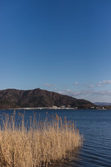 Vue sur le lac Kawaguchi. On voit le lac, au loin un pont et quelques maisons. Tout au fond des petites montagnes ou grandes collines. 