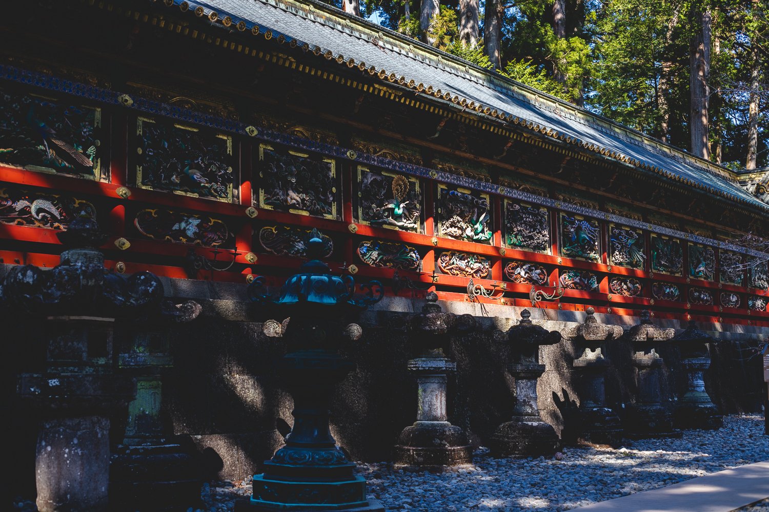 Le mur d'un batiment du sanctuaire de Nikko. Le tout est couvert de bois sculpté et peint formant des bas reliefs de couleurs vives. Devant le mur se trouve une rangée de lanternes de pierre.