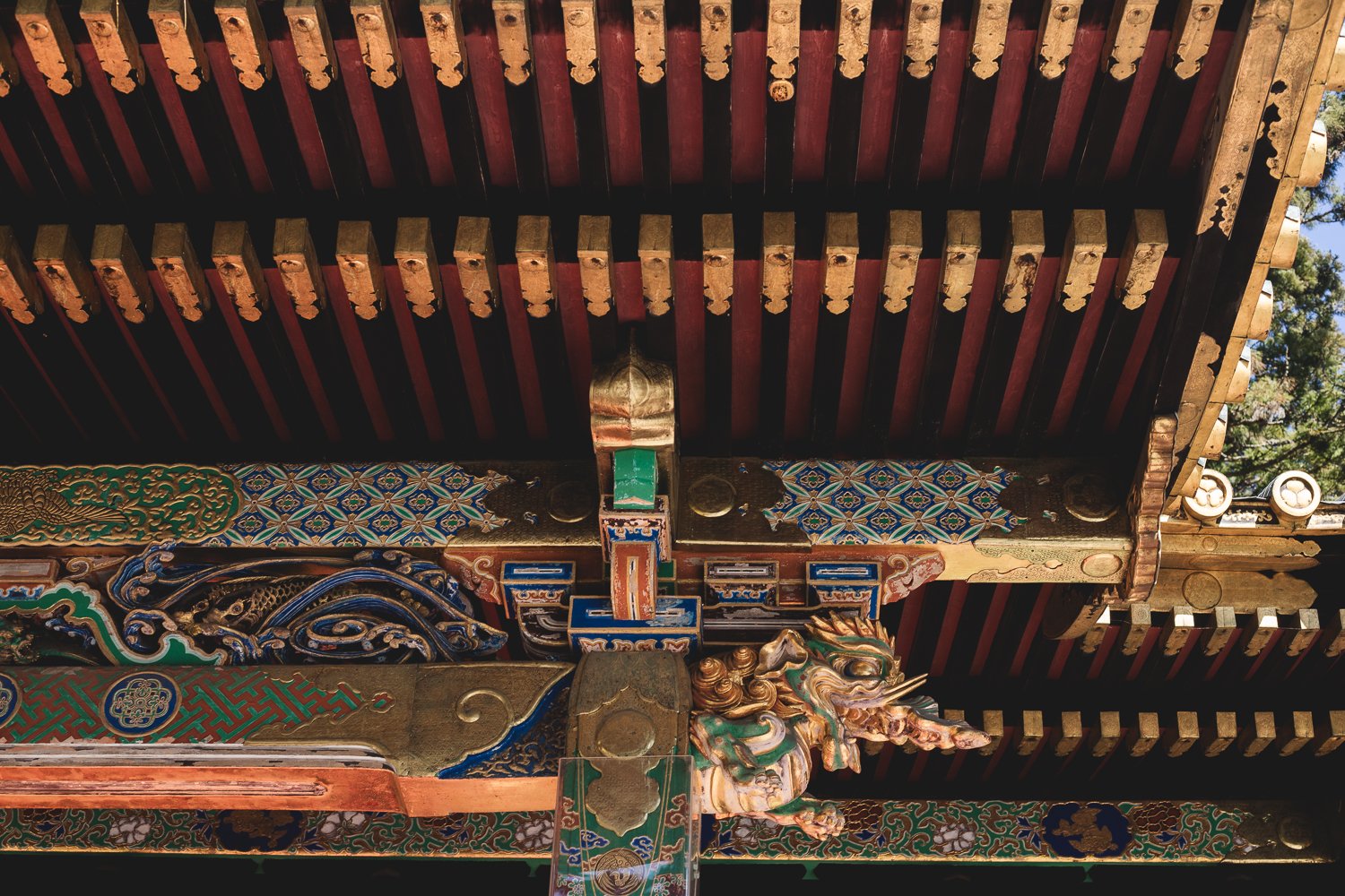 Détail d'une sous-toiture d'un bâtiment du sanctuaire. Les poutres sont couvertes de motifs géométriques peints de couleurs vives. Une d'elle se termine en formant une tête de dragon sculptée.