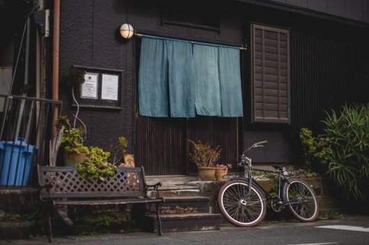 Un vélo est posé devant une maison, on en voit la porte en bois masquée par des tissus bleus. Devant la maison se trouve un banc. La scène est pittoresque et reflète bien l'ambiance des petites rues annexes à Kamakura