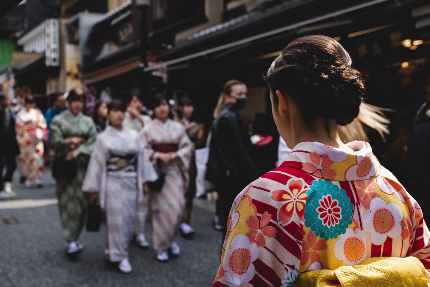 Une personne marchant dans la rue,de dos et portant un yukata. En face dans le flou de la photo, on devine d'autres personnes vêtues de Yukata et avançant en sens inverse